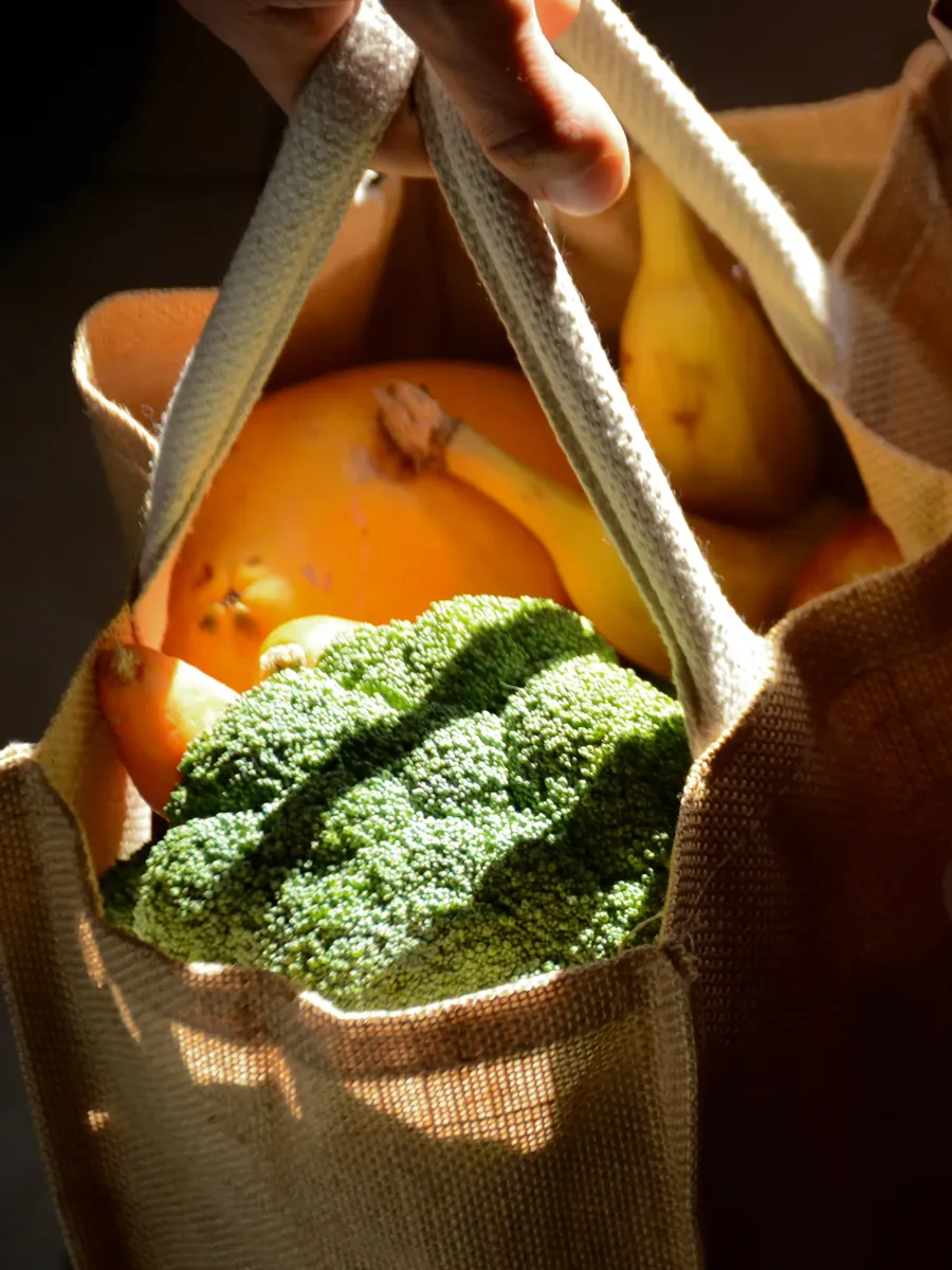 Una bolsa de mandado con brócoli, papaya y plátanos, sostenida a contraluz